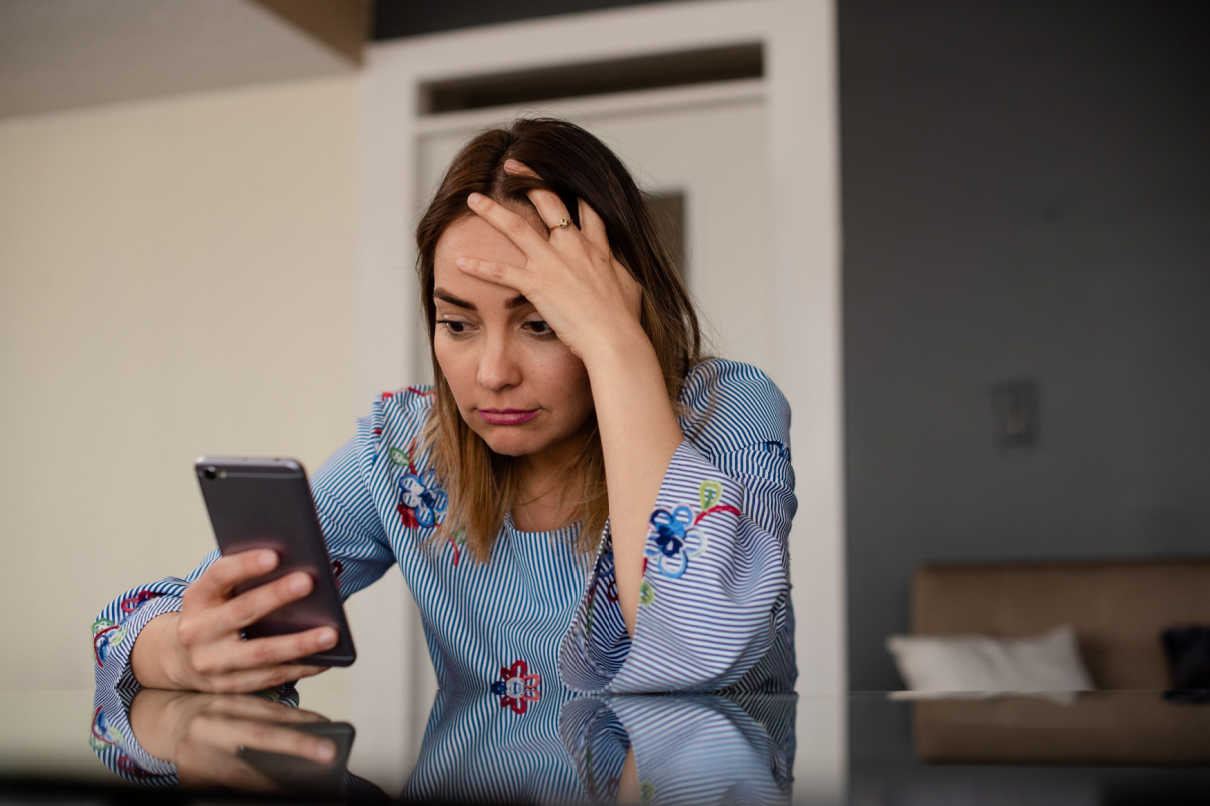 Imagem de mulher com a mão na cabeça, sinalizando indignação, enquanto olha para o celular